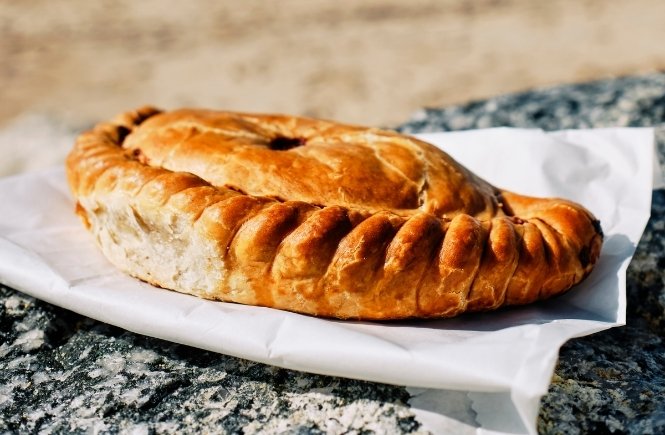 A Cornish pasty resting on a wall in front of a beach on a sunny day