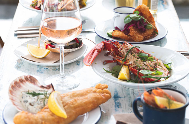 A table full of various seafood dishes and glasses of white wine at Mackerel Sky Seafood Bar