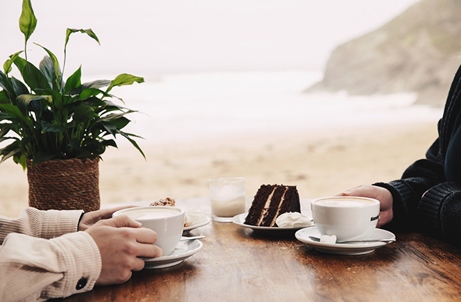 Two people enjoying a coffee and a cake at Lusty Glaze with the beach behind them
