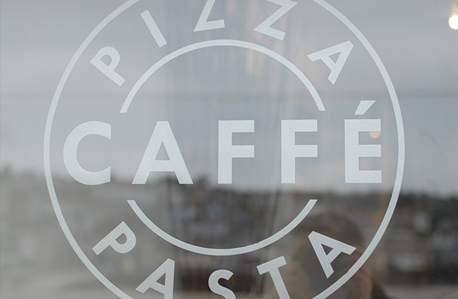 The sign of Caffe Pasta on the window of their shop with the harbour reflected in the glass