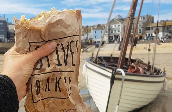 Someone holding up a St Ives Bakery Cornish pasty on Harbour beach in St Ives with a boat in the background