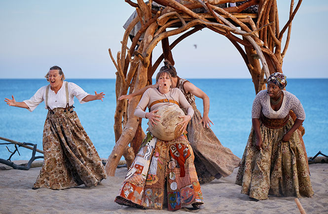 Actors dancing in a ring of wood on the beach at Carlyon Bay with a wooden tree house behind them in Wildworks' production of I AM KEVIN