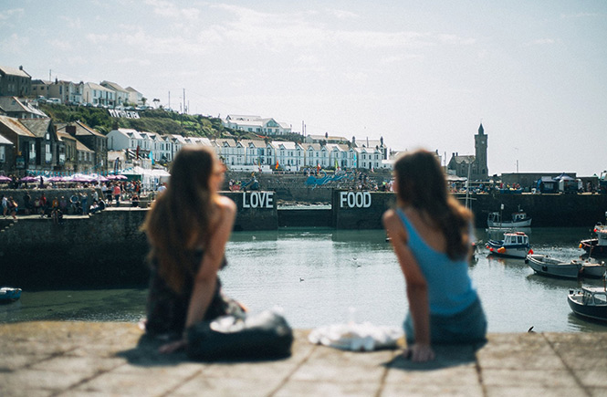 Huge white letters saying Love Food sit above the harbour of Porthleven during the Porthleven Food Festival as two women sit and eat by the water