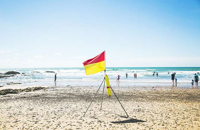 A red and yellow flag flutters in the sea breeze whild people splash in the sea.