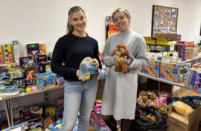 Two people hold toys, surrounded by hundreds of other donated toys.