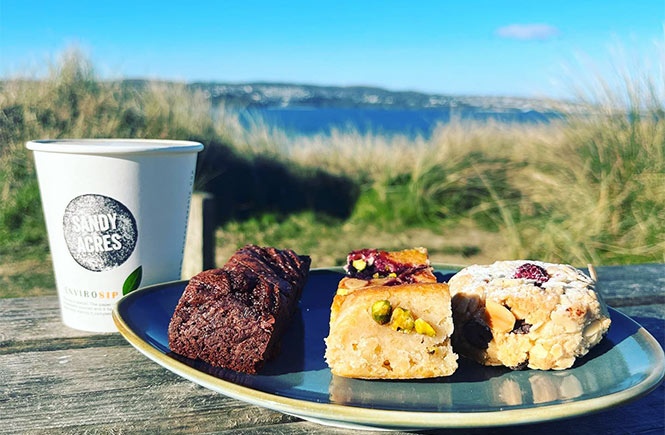 Pieces of cake on a plate with a takeaway coffee overlooking sand dunes and the sea at Sandy Acres Beach Café in Cornwall