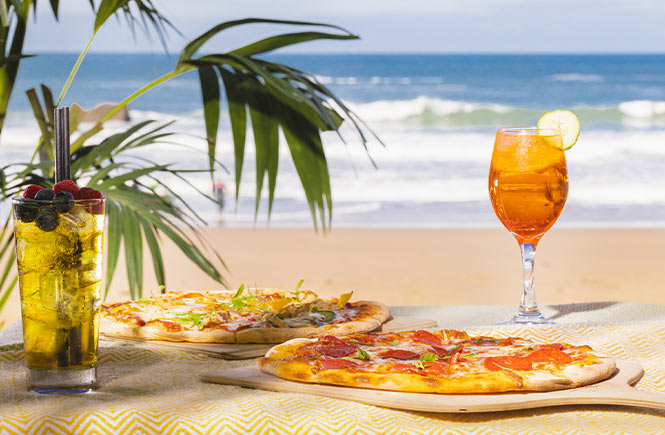 Two pizzas and cocktails with the beach and sea in the background at Lusty Glaze Restaurant in Newquay