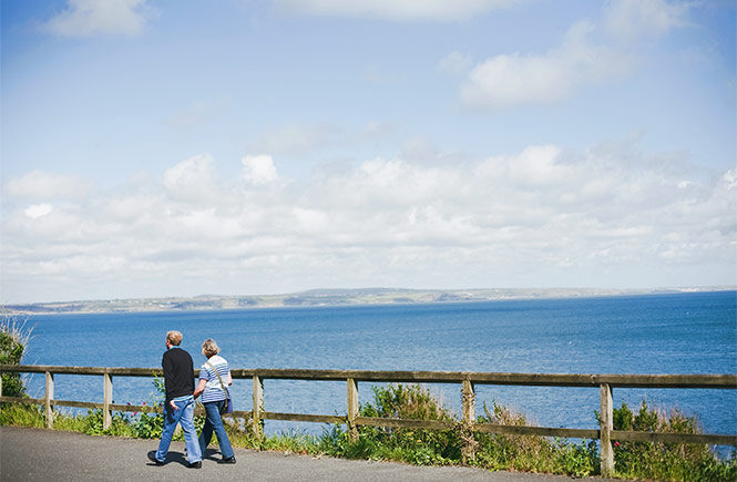 Two people walking along a flat and accessible path by the water between Newlyn and Mousehole in Cornwall
